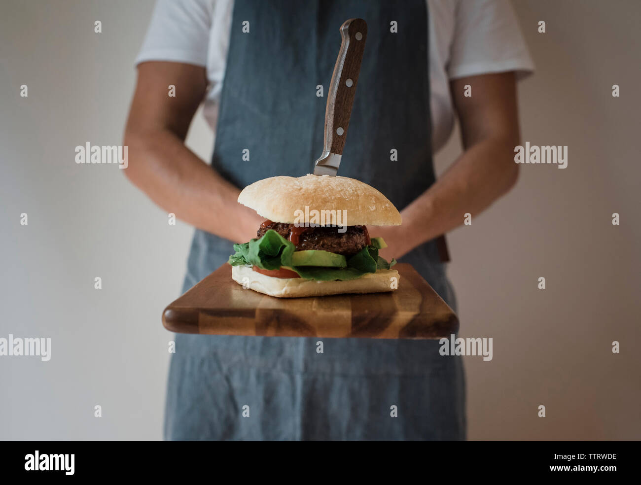 Mittelteil der Mann hält Burger mit Messer auf Schneidebrett zu Hause Stockfoto