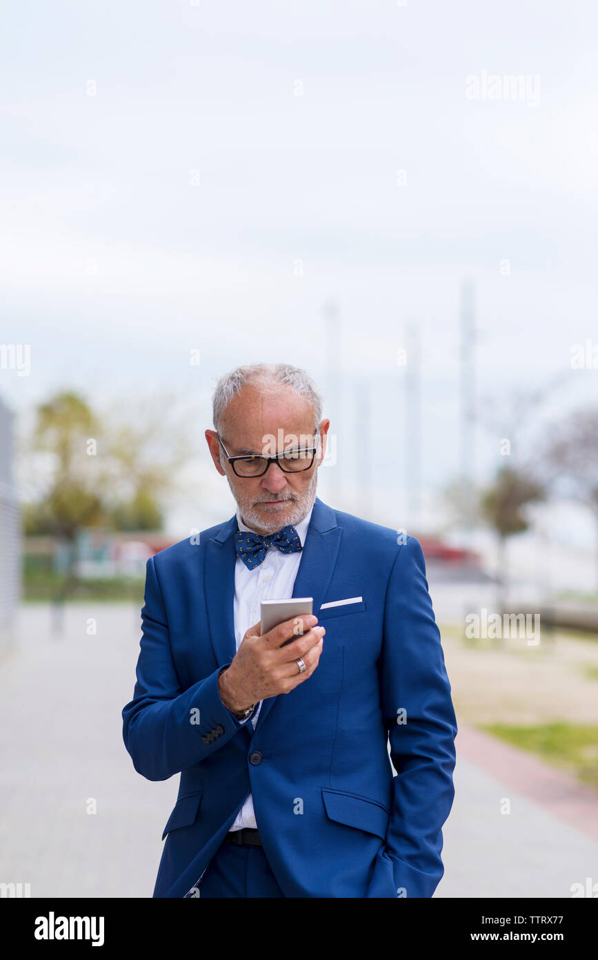 Zuversichtlich älterer Mann im Anzug mit Smart Phone beim Stehen auf Fußweg gegen Sky Stockfoto