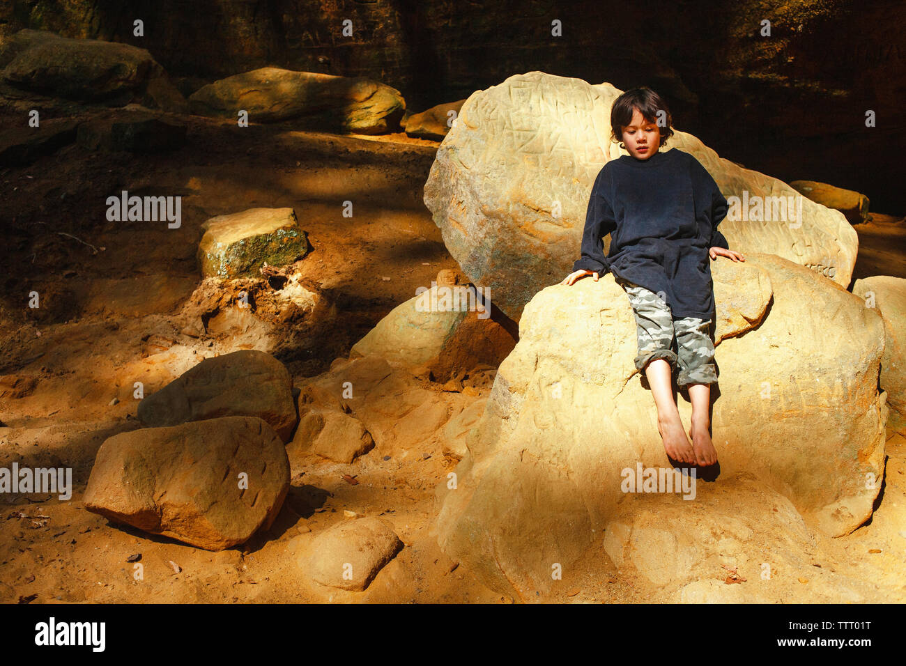 Ein kleiner Junge sitzt barfuß in einem Pool von goldenem Licht in einer felsigen Schlucht Stockfoto