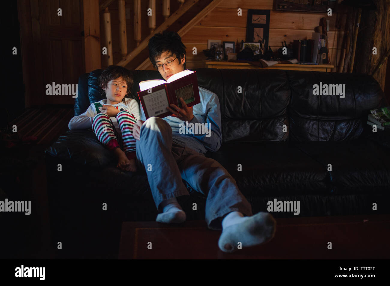 Ein kleiner Junge im Schlafanzug sitzt mit Vater, der in den frühen Morgen lesen Buch Stockfoto