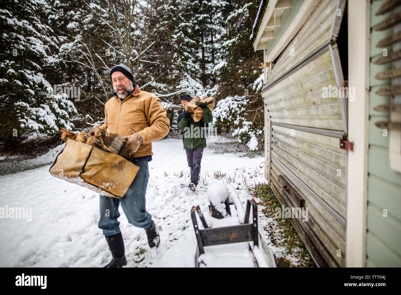 Zwei Männer tragen Holz für Kamin an einem verschneiten Wintertag durch eine Garage Stockfoto