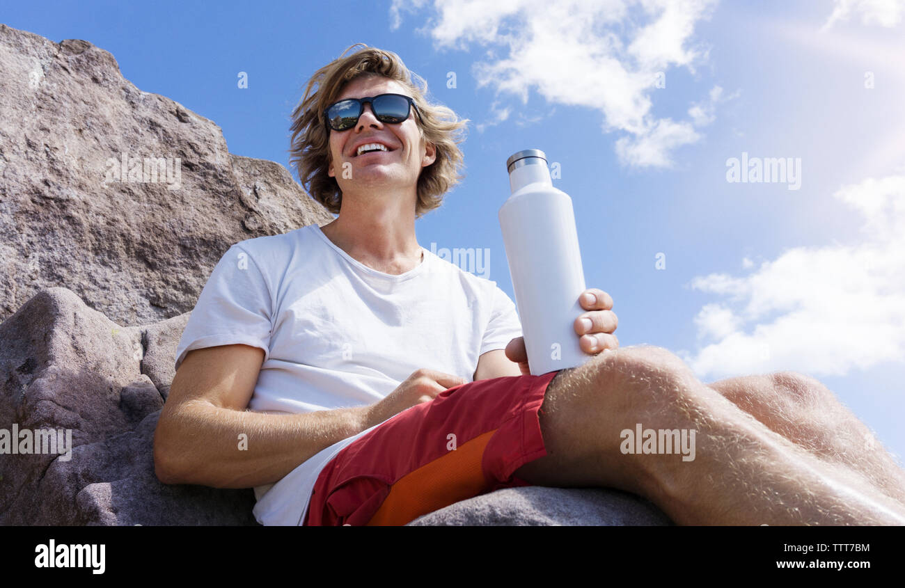 Low Angle View der glücklichen Mann hält die Flasche beim Sitzen auf Rock Formation gegen Sky Stockfoto