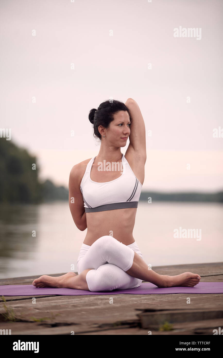 Selbstbewusste Frau üben Kuh Gesicht Pose auf Pier am See gegen Himmel bei Sonnenuntergang Stockfoto