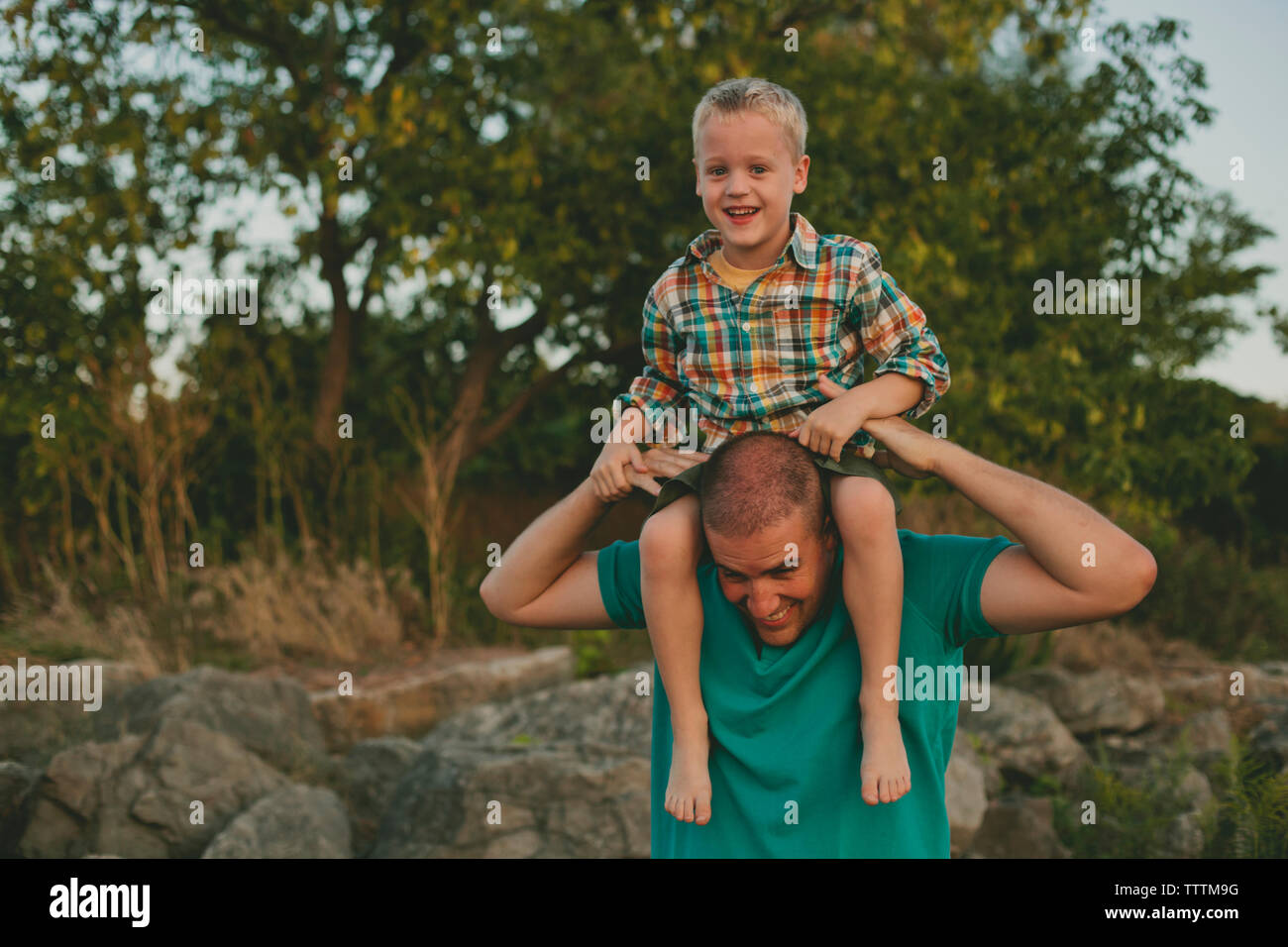 Gerne Vater mit Sohn auf den Schultern stehend gegen Bäume Stockfoto
