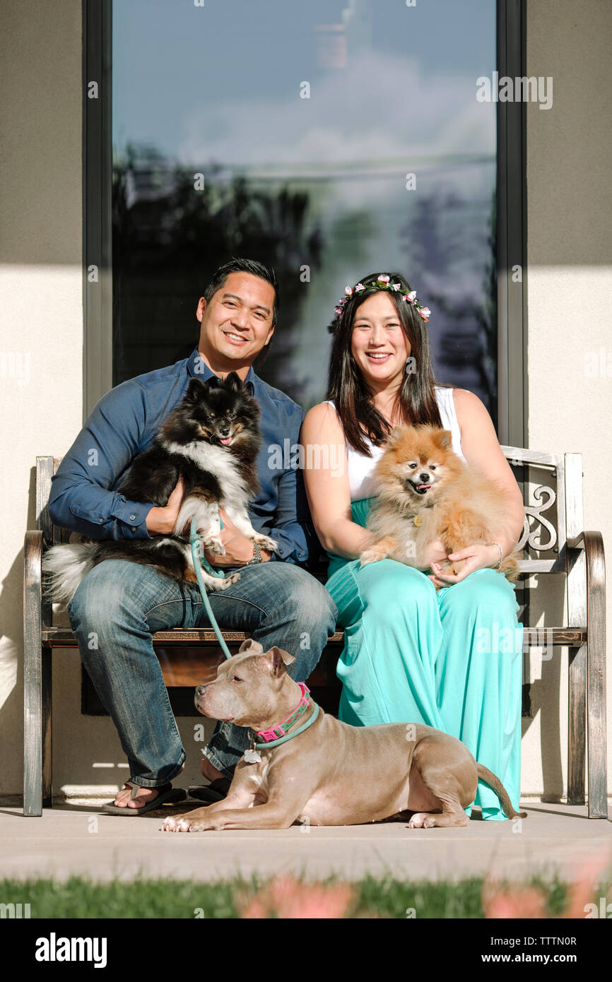 Portrait von zuversichtlich lächelndes Paar mit Hund sitzt auf der Bank gegen Haus Stockfoto