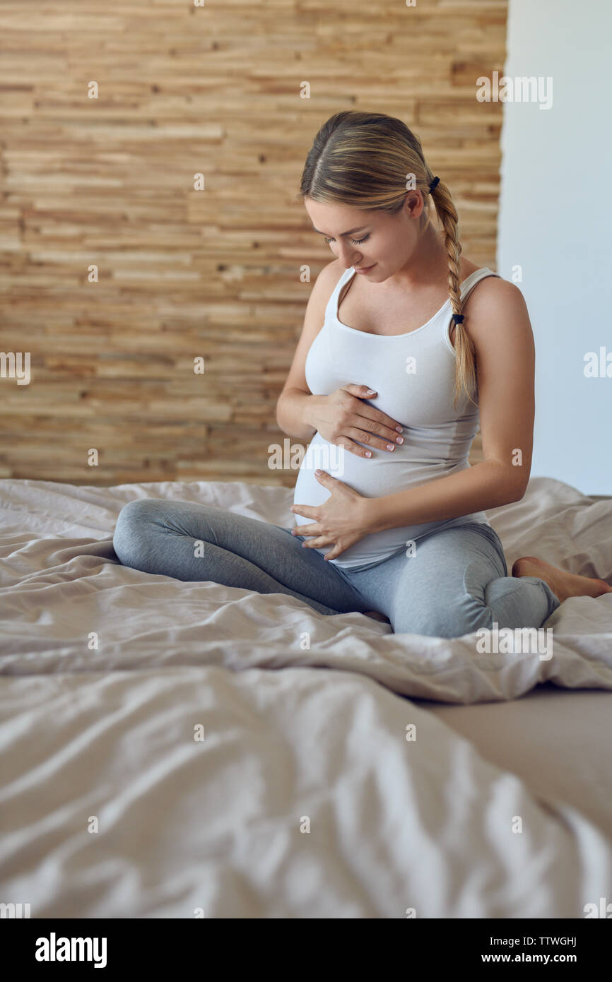 Glückliche junge schwangere Frau Verklebung mit ihrem ungeborenen Kind sitzen auf einem Bett mit Ihrem Babybauch mit ihren Händen, mit einem zarten Lächeln der Lo Stockfoto