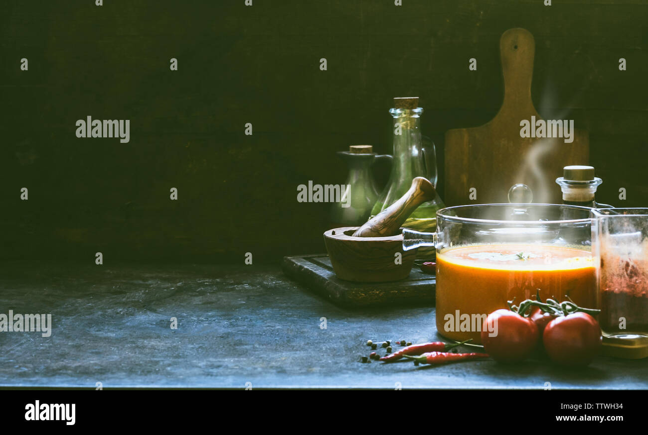 Heiße Tomatensuppe mit Dampf in Glas Topf auf rustikalen Tisch bei dunkler Hintergrund mit Zutaten zum Kochen. Küche Szene, noch Leben. Hausgemachte gesund Stockfoto