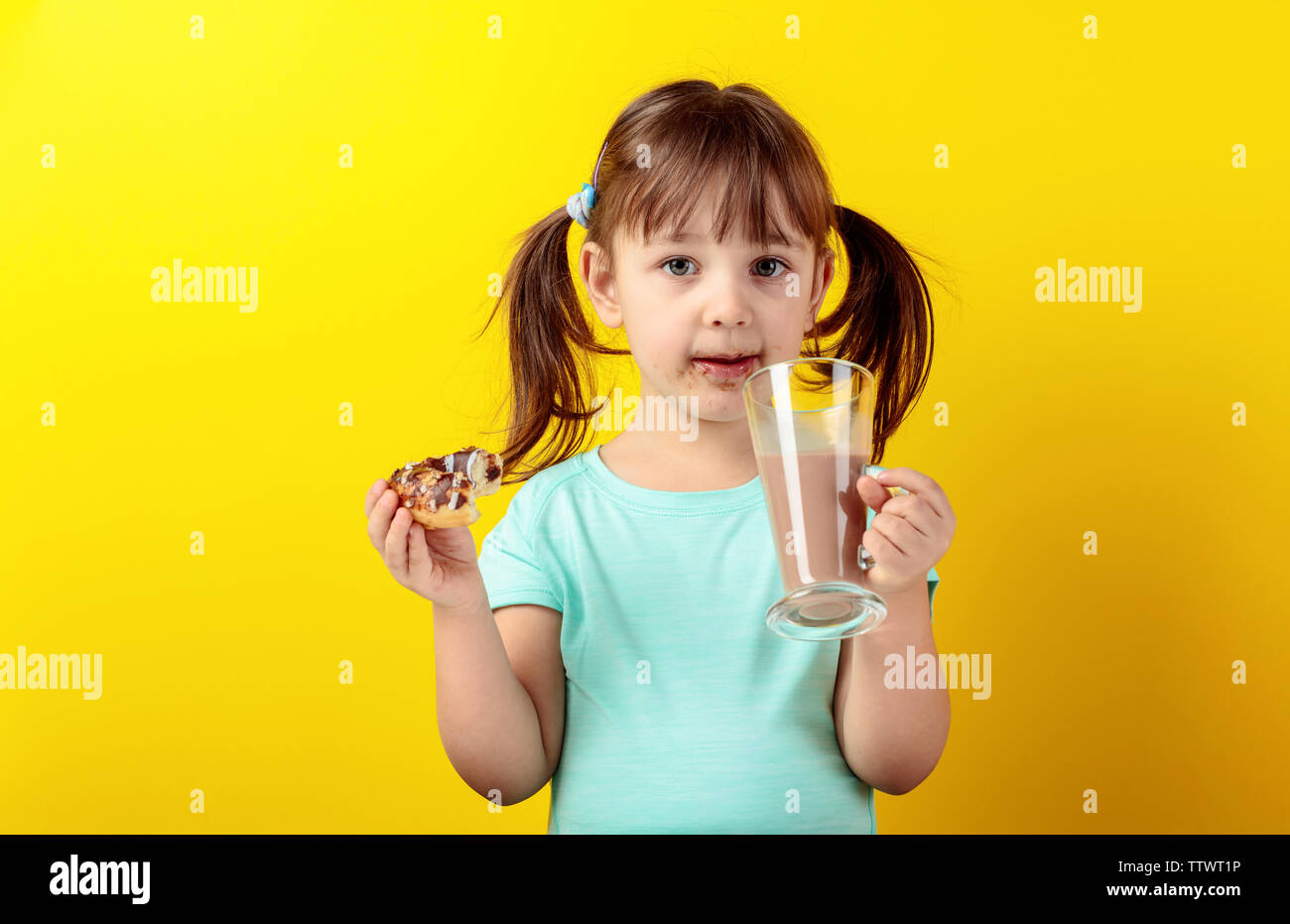 Kleines Mädchen in einem Türkis t-shirt Donuts essen und trinken Kakao. Die Haare des Mädchens ist in Schwänze gebunden. Gelber Hintergrund. Stockfoto