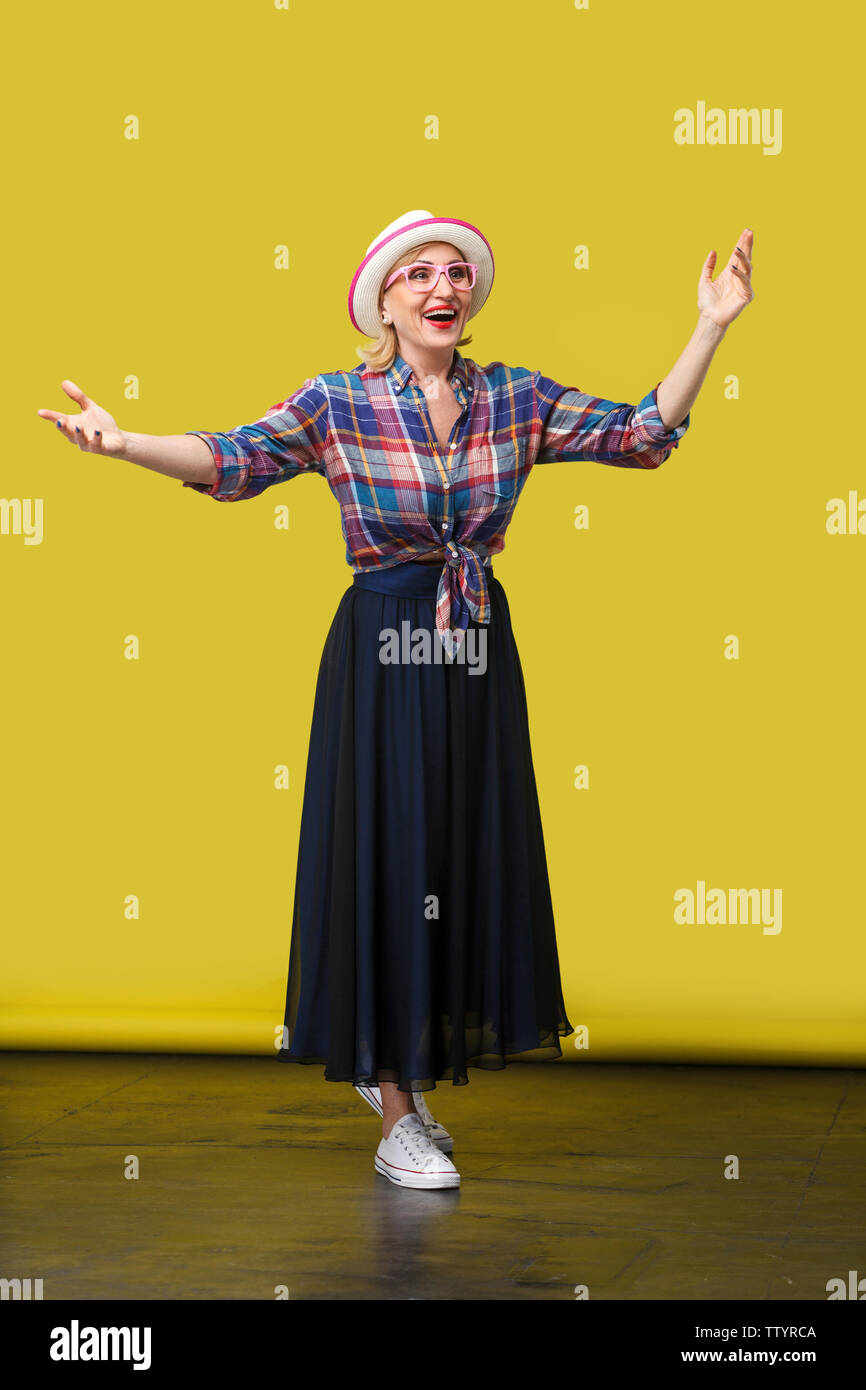 Volle Länge Portrait von glücklich moderne, elegante, reife Frau im lässigen Stil mit Hut und Brille stehend mit erhobenen Armen und überraschte Gesicht Stockfoto