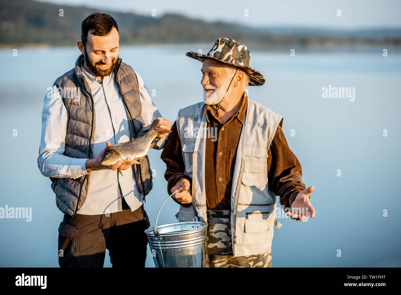 Gerne Großvater mit erwachsenen Sonne stehen zusammen mit frisch gefangenen Fisch auf der Pier in der Nähe des Sees in den frühen Morgenstunden Stockfoto