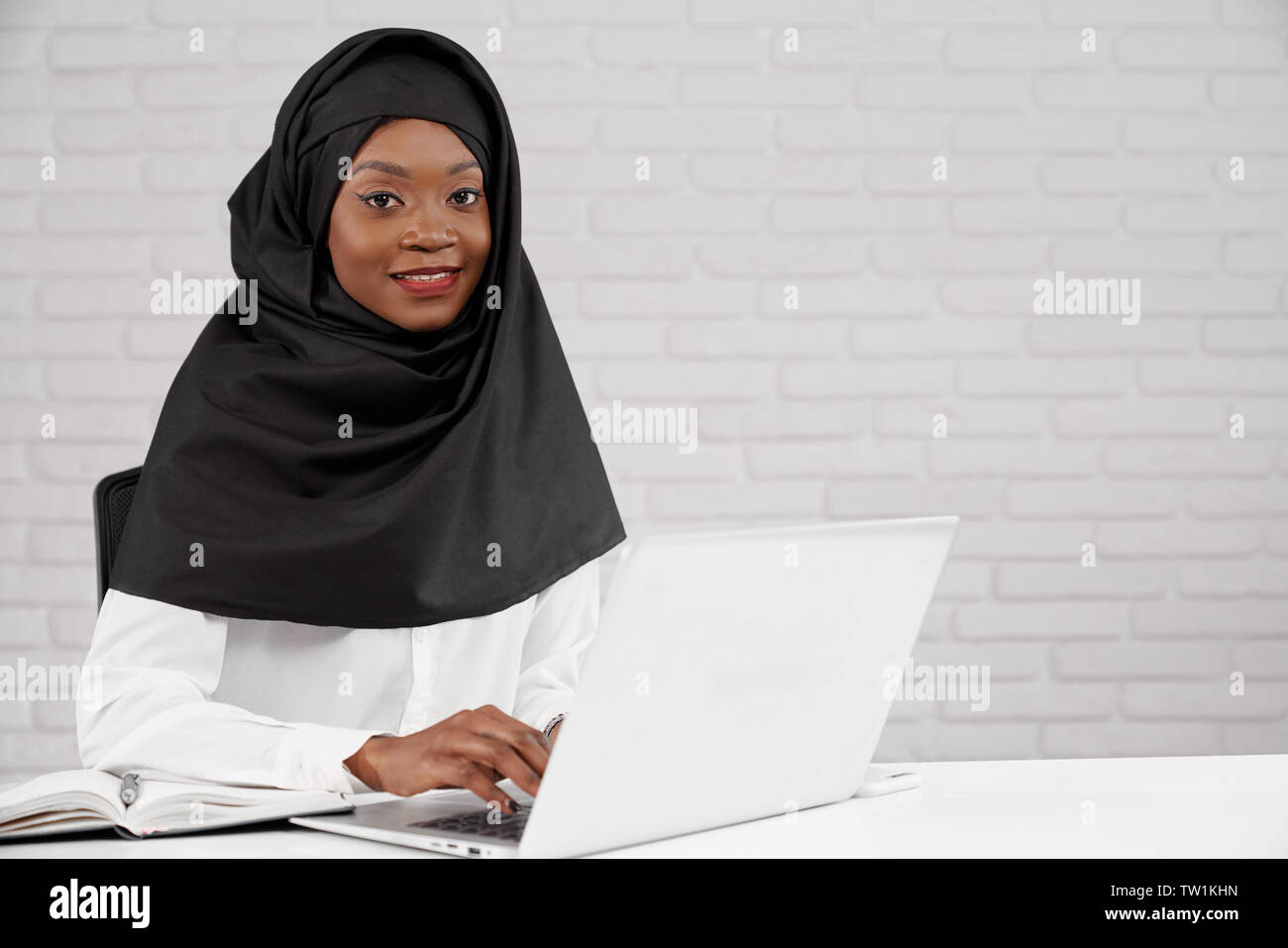 Hübsches, stilvolles, jungen afrikanischen Mädchen im Büro sitzen, mit Laptop. Schöne muslimische Frau mit Kopftuch in schwarze und weiße Kleidung lächelnd in die Kamera schauen. Stockfoto