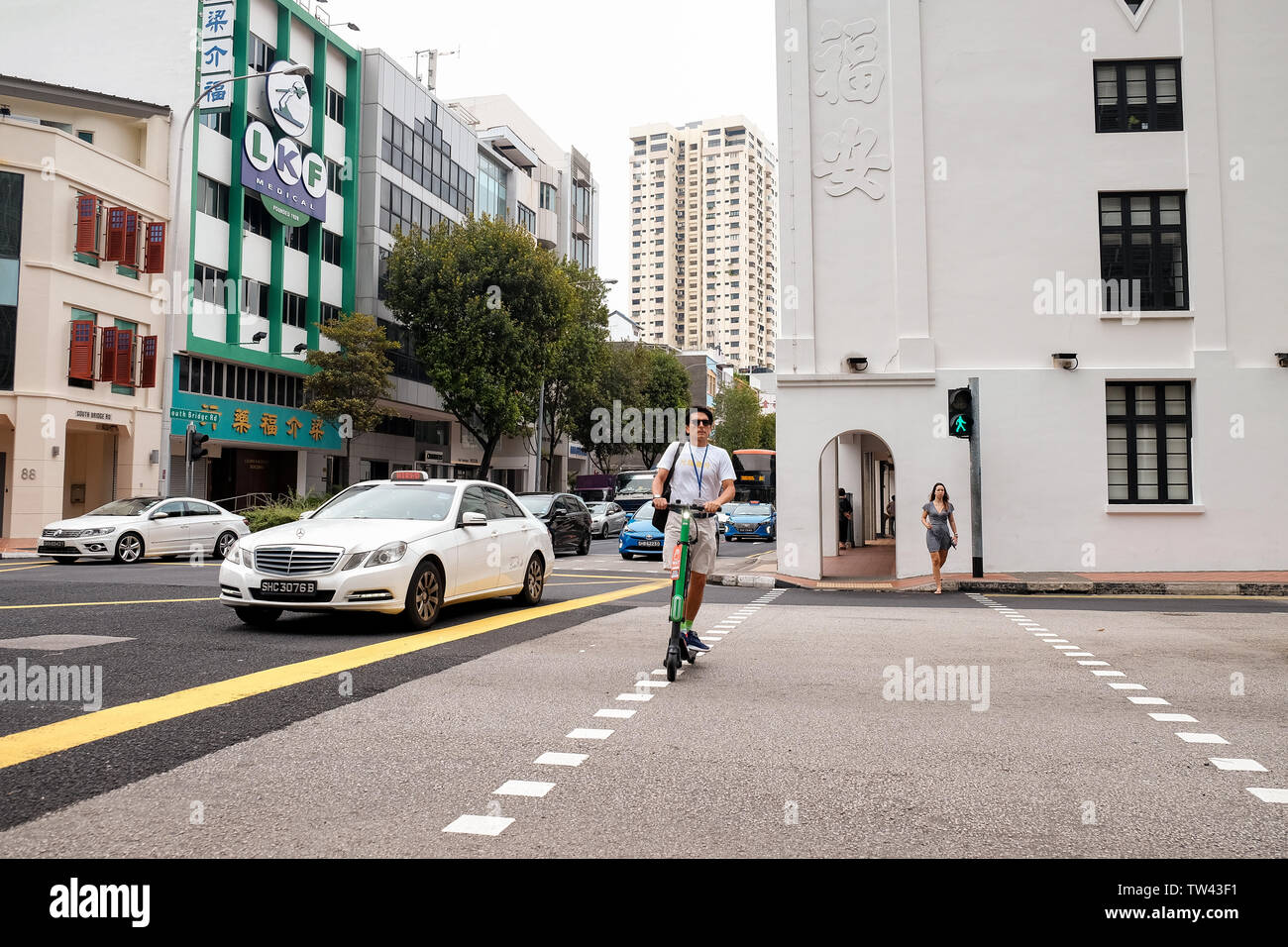 Ein alter Mann in Shorts, T-Shirt und Sonnenbrille verwendet einen elektronischen Roller eine Kreuzung in Singapur zu überqueren. Stockfoto