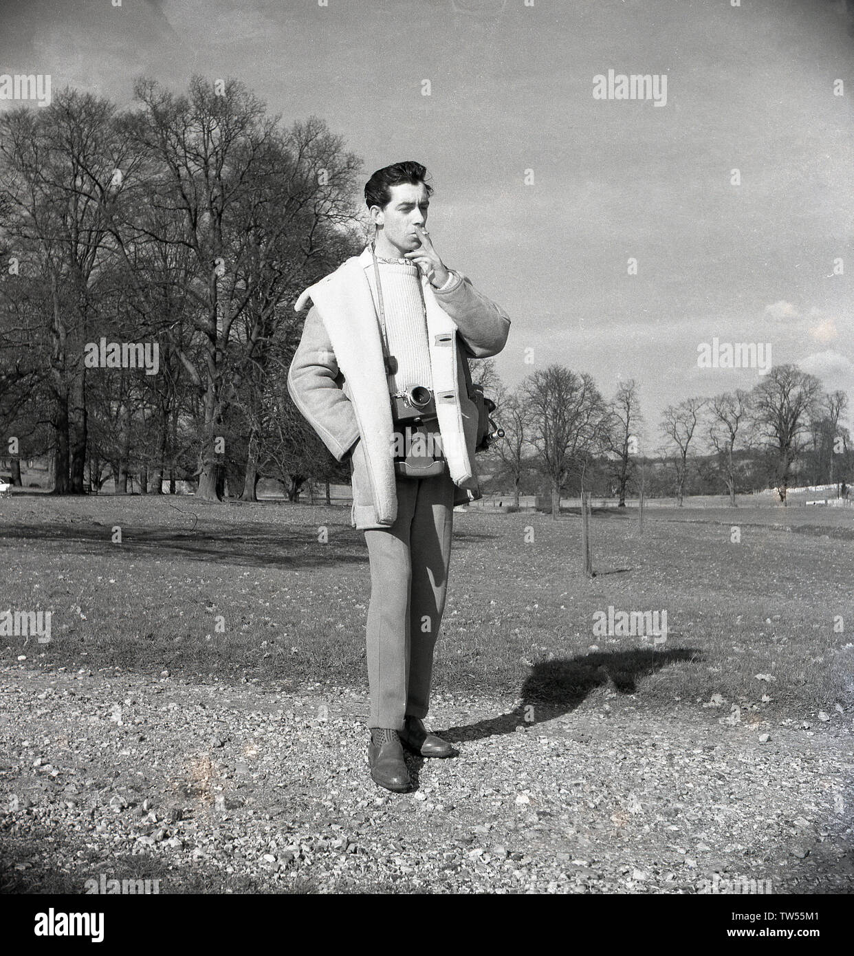 1960, historische, Fotograf... ein Junge bin ein über Stadt' mit seiner Filmkamera, draußen auf einem Schotterweg in einem Park in den Moden des Tages, das Tragen eines crewneck Wollpullover, eine Krawatte und einem dicken Schaffell Jacke und rauchen eine Zigarette, England, UK. Stockfoto