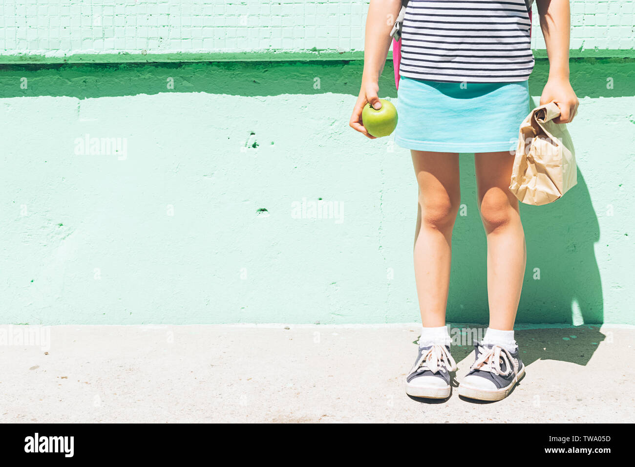 Ein kleines Mädchen in Sneakers hält einen Apfel und eine Papiertüte mit einer Schule Snack vor dem Hintergrund einer grünen Wand. Stockfoto