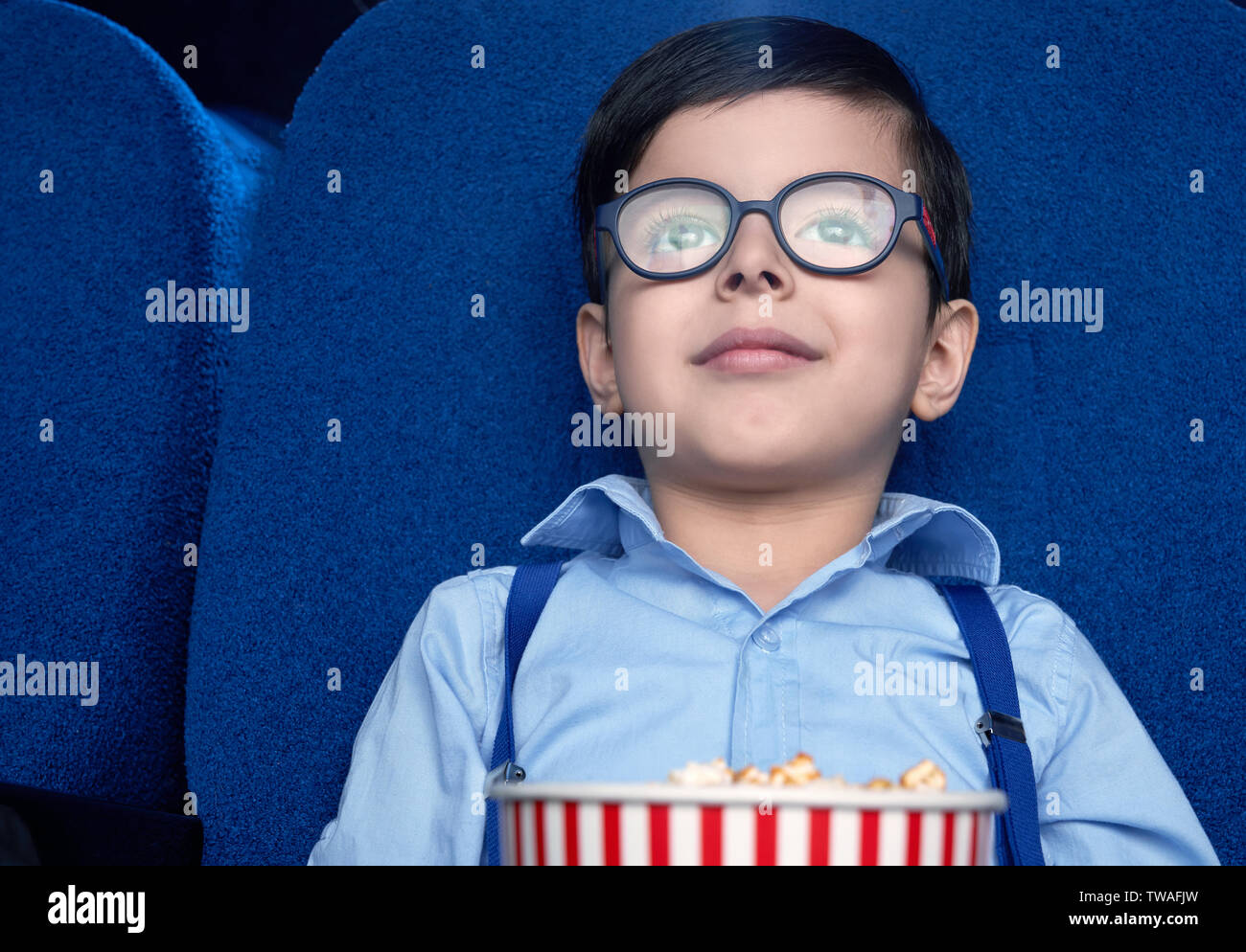 Vorderansicht des lustigen, kleinen Jungen im Kino sitzen und beobachtete aufgeregt Film. Junge Zuschauer Brillen und weißes Hemd lächelnd und Entspannung am Wochenende. Konzept der Unterhaltung und des Genusses. Stockfoto