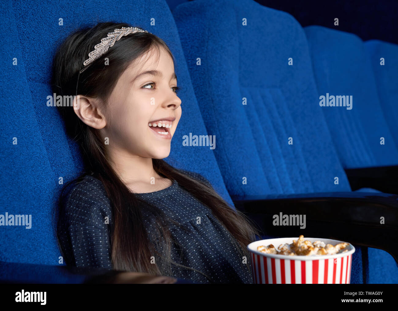 Seitenansicht der kleine Dunkelhaarige sitzen auf Blau Sitz im Kino, Komik und Lachen. Fröhliches schönes Mädchen Popcorn essen und genießen lustiger Film im Kino. Konzept der Unterhaltung. Stockfoto