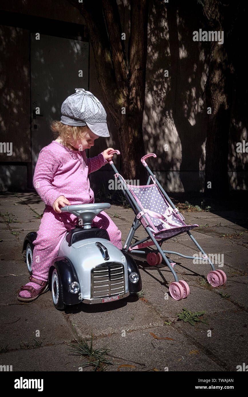 Kleines Kind spielen im Freien an einem sonnigen Sommertag, in ihrem rosa Schlafanzug, sitzen auf ein Spielzeugauto, ein Puppenwagen Stockfoto