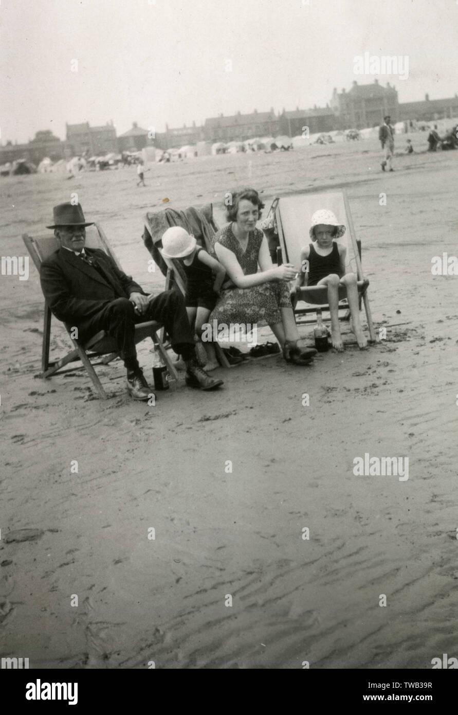 Familie Gruppenfoto am Meer. Ein älteres Paar sitzen auf den Liegestühlen mit zwei Mädchen in die passende Hüte und Badesachen, die Erwachsenen lächelnd, während die Kinder schauen weniger sicher das theyre, das sich zu amüsieren. Der Vater oder Großvater trägt robuste Stiefel und hat eine Flasche trinken unter seinem Liegestuhl! Datum: ca. 1920 s Stockfoto