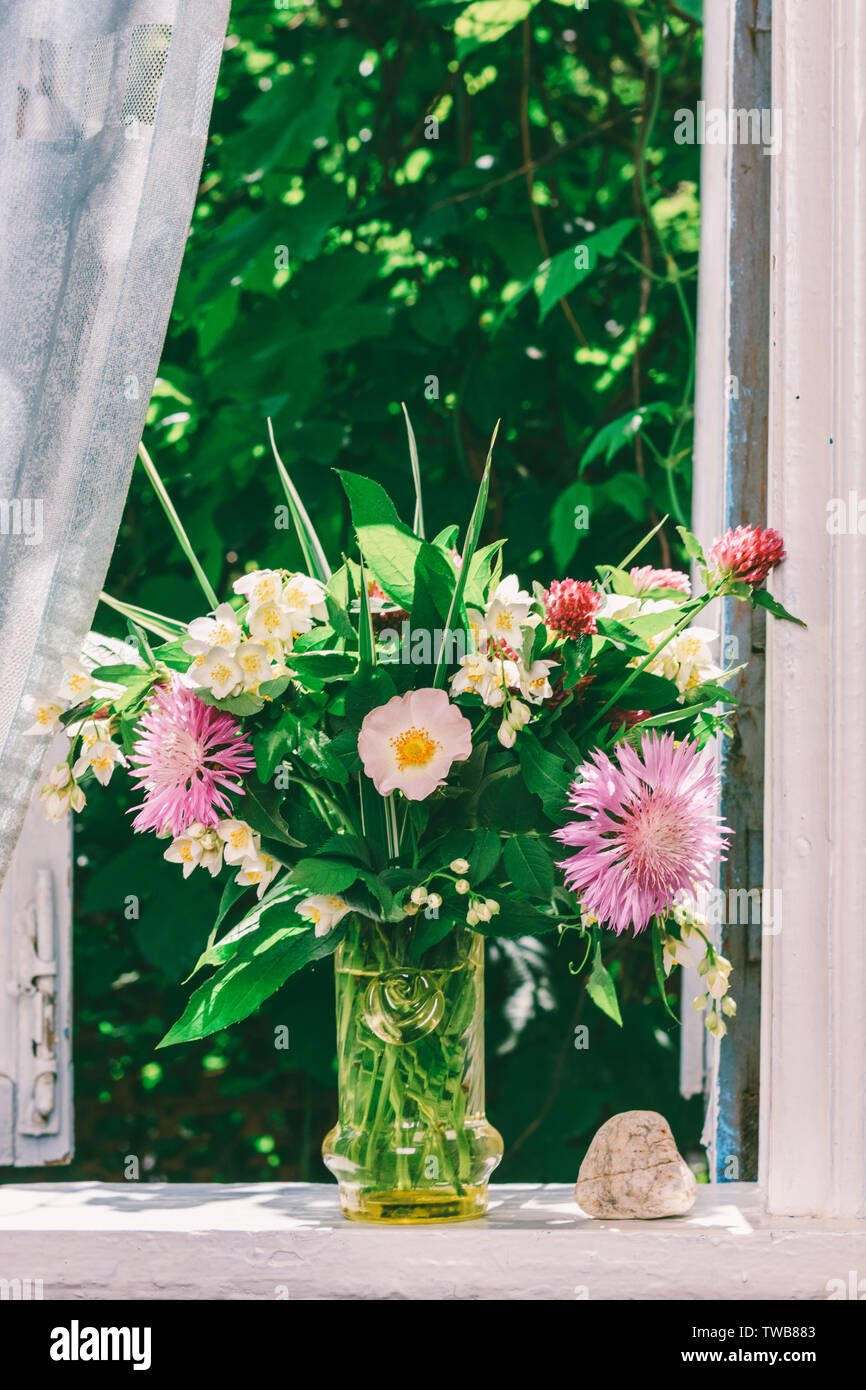 Ein Blumenstrauß von Clover, Kornblumen und Jasmin in einem Glas Vase und ein Herz aus Stein auf der Fensterbank eines offenen Fensters eines Dorfes Haus Stockfoto
