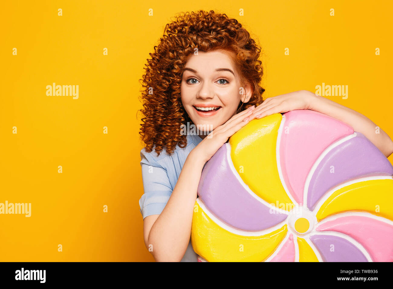 Curly Rothaarige positive Frau trägt blaue Kleid mit süßen großen Lollipop auf gelbem Hintergrund Stockfoto