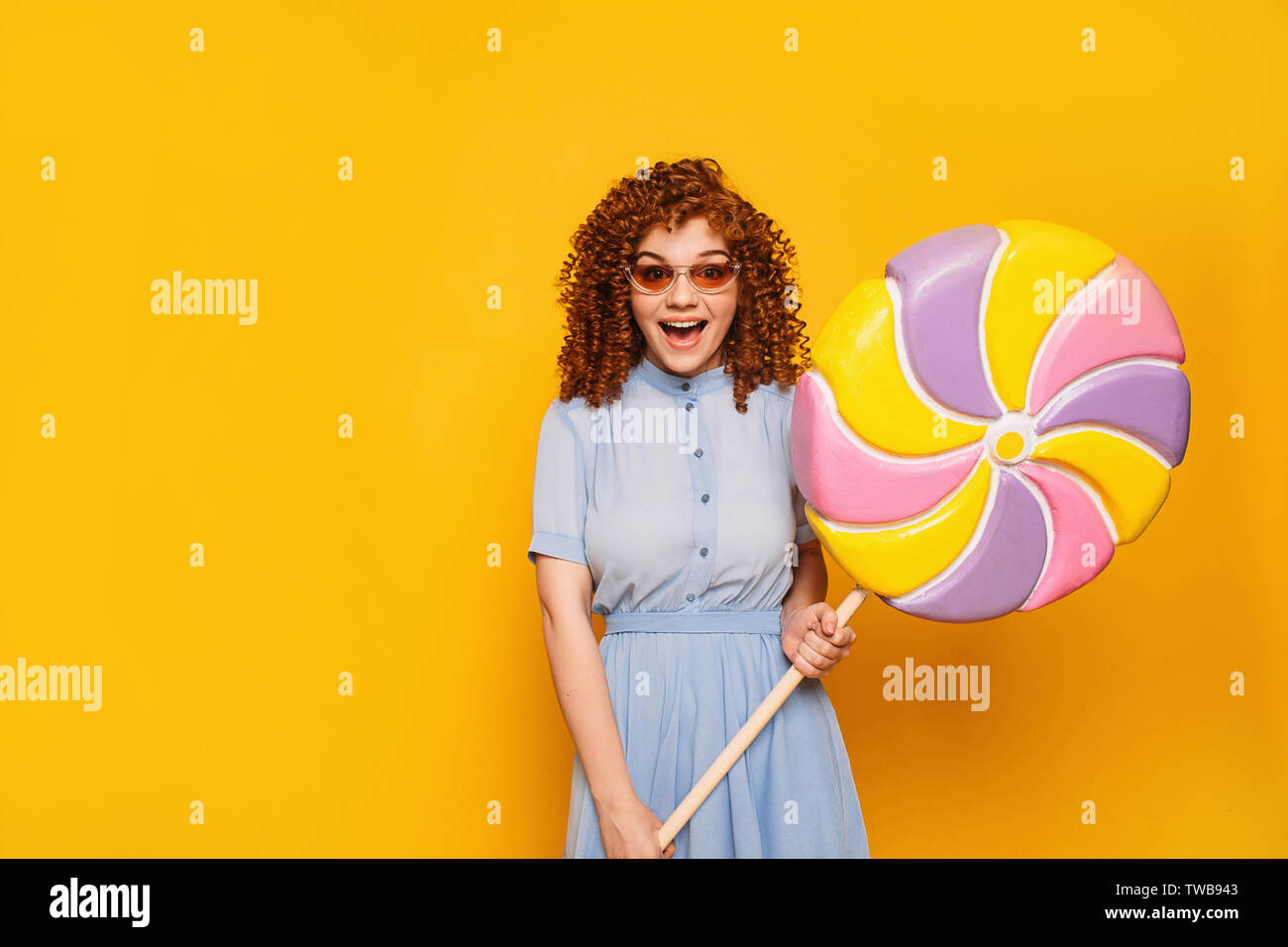 Curly Rothaarige positive Frau trägt blaue Kleid mit süßen großen Lollipop auf gelbem Hintergrund Stockfoto