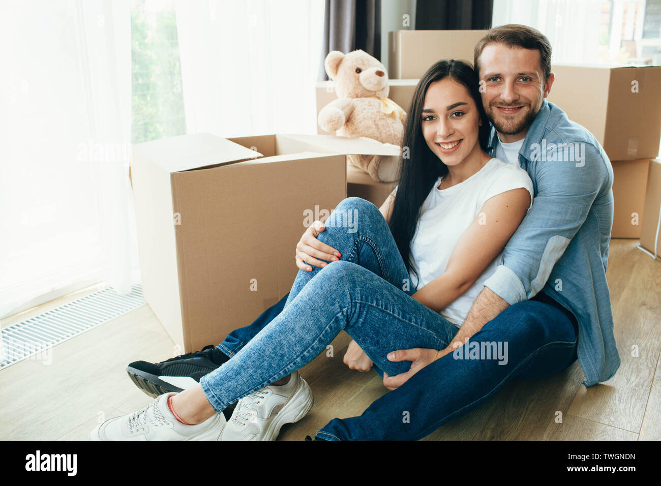 Glückliches Paar umarmen und sitzen auf dem Boden in Ihrem neuen Zuhause mit vielen Kartons auf Hintergrund Stockfoto