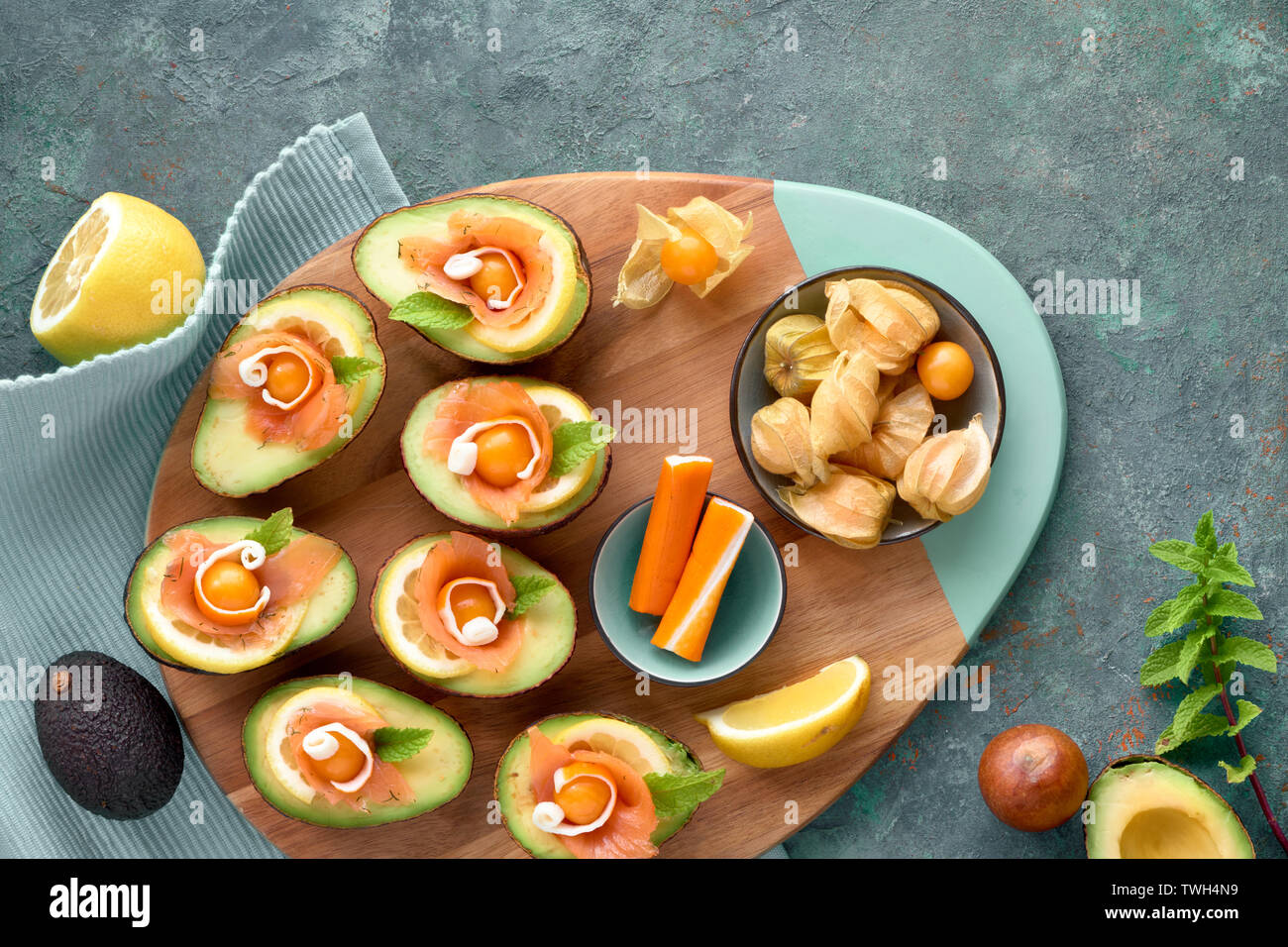 Avocado Boote mit Räucherlachs, Crab Sticks, Zitrone, Physalis und Minzeblatt, Ansicht von oben auf der cyanfarbenen strukturierten Hintergrund Stockfoto