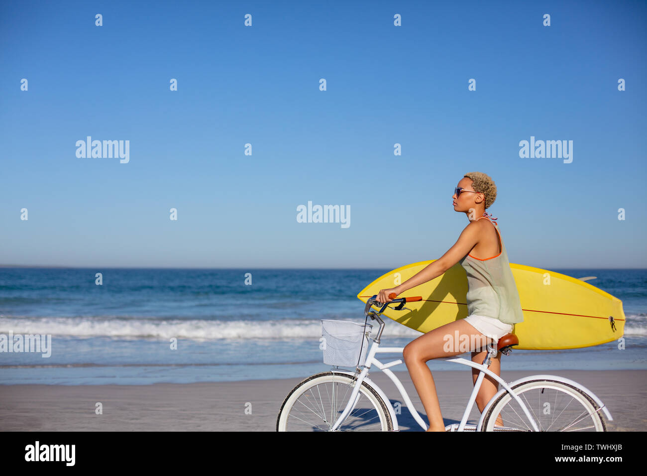 Frau mit Surfboard sitzen auf dem Fahrrad am Strand in der Sonne Stockfoto