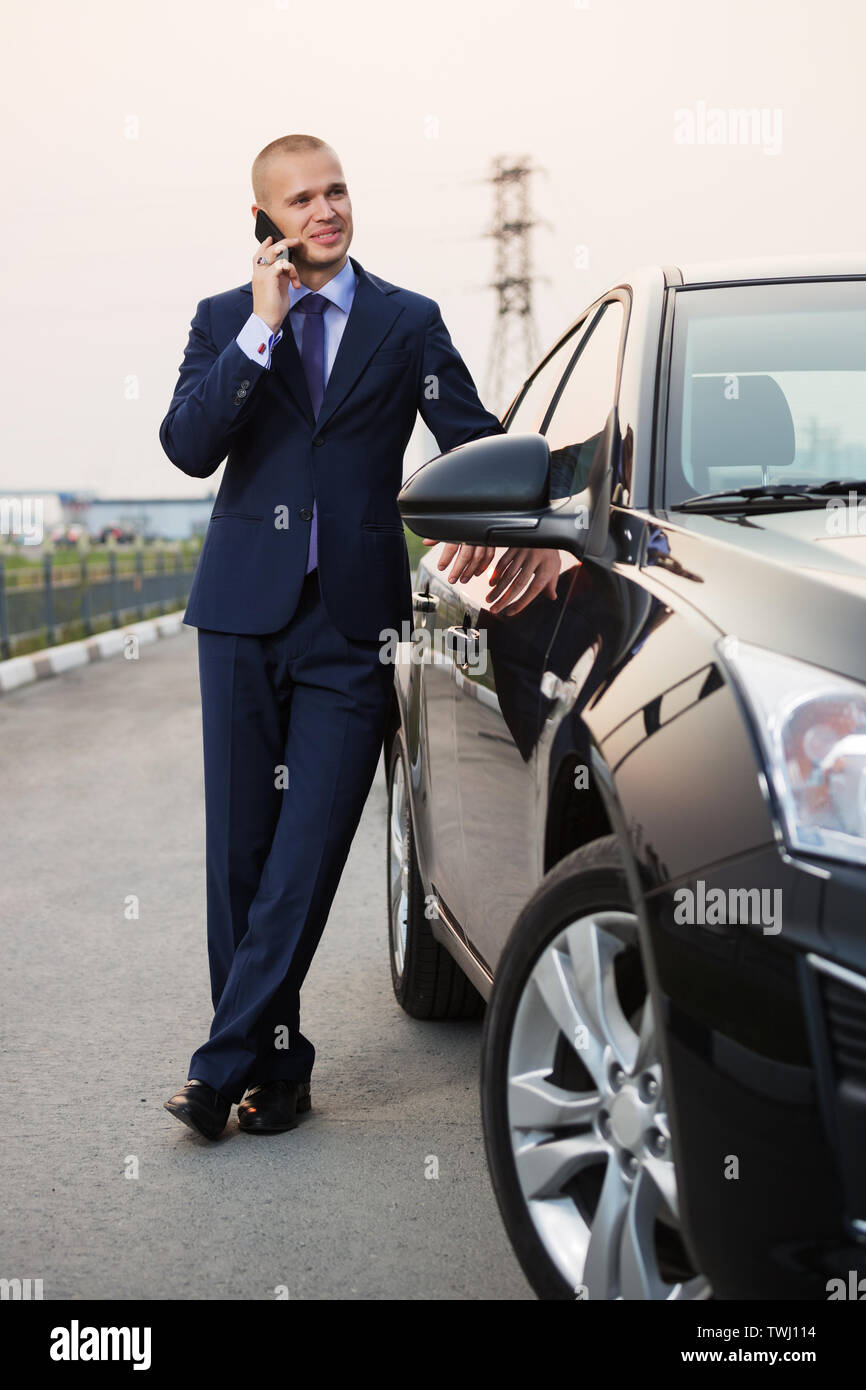 Junge Unternehmer, die auf dem Mobiltelefon neben seinem Auto stilvoll trendy männliches Modell tragen dunkelblauen Anzug Stockfoto