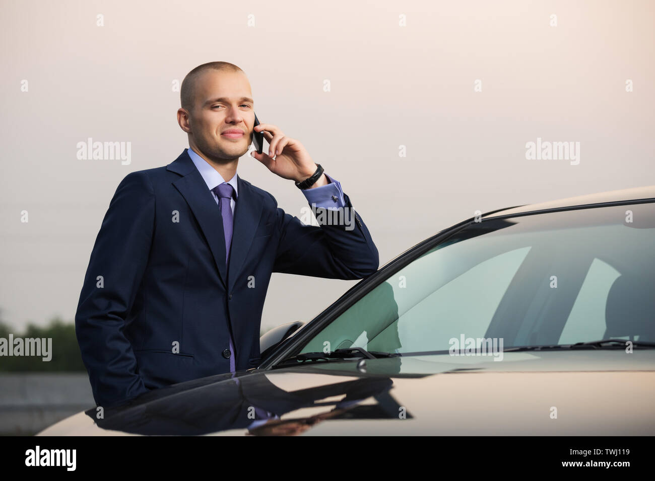 Junge Unternehmer, die auf Handy lehnte sich auf seinem Auto stilvoll trendy männliches Modell tragen dunkelblauen Anzug Stockfoto