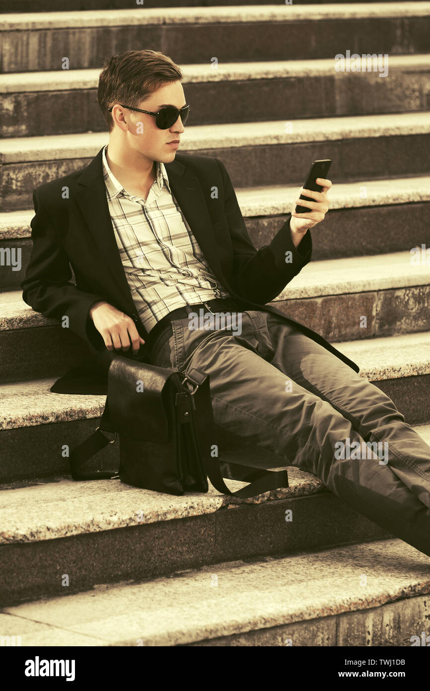 Young Business Mann mit Handy sitzen auf Schritte Stilvolle männliches Modell in Schwarz Blazer und Hose grau Stockfoto