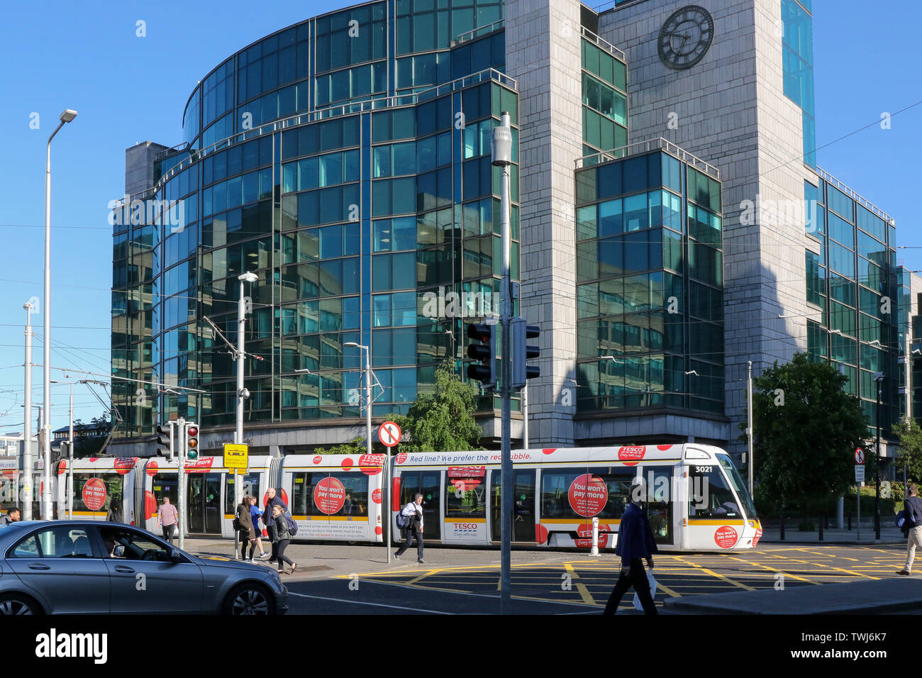 Menschen zu Fuß in einem Dubliner Straße an einem sonnigen Sommerabend mit einer Straßenbahn Luas in Dublin vor ein modernes Gebäude mit einer Glasfassade. Stockfoto