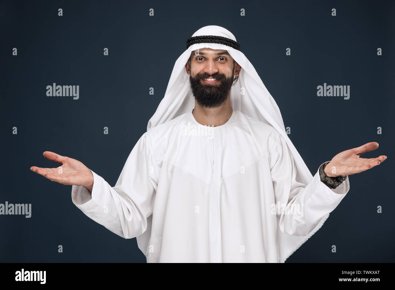 Brustbild des Arabischen saudischen Geschäftsmann auf dunkelblau studio Hintergrund. Junge männliche Modell Lächeln, mit einer Geste der einladend. Konzept der Business, Finanzen, Mimik, menschliche Gefühle. Stockfoto