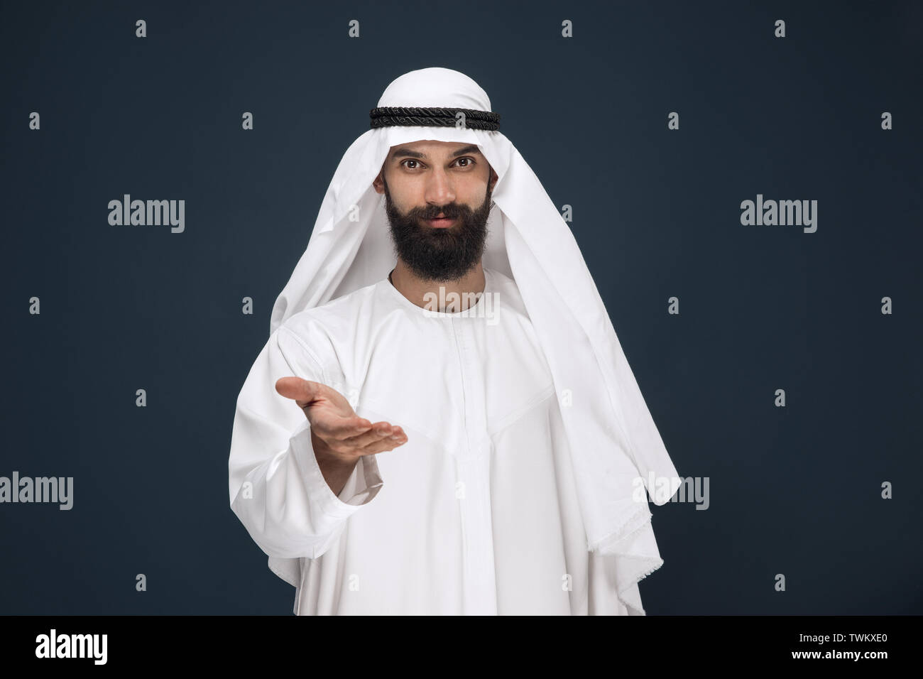 Brustbild des Arabischen saudischen Geschäftsmann auf dunkelblau studio Hintergrund. Junge männliche Modell Lächeln, mit einer Geste der einladend. Konzept der Business, Finanzen, Mimik, menschliche Gefühle. Stockfoto