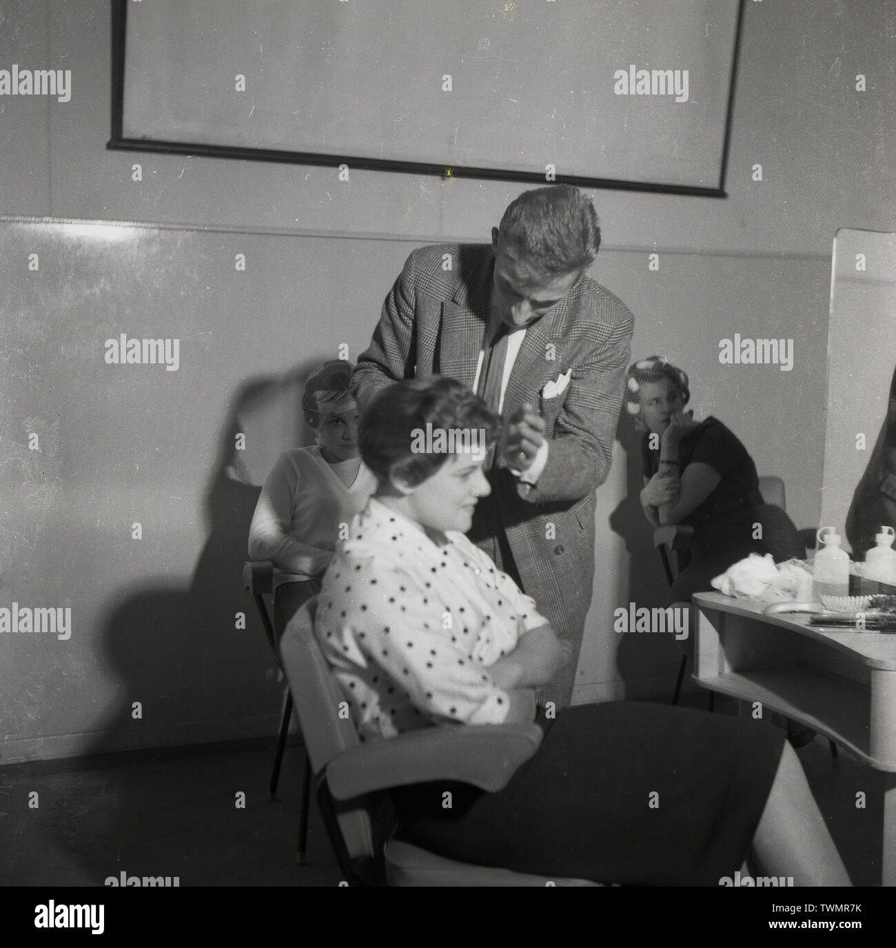 1960, historische, bei Friseur Schule oder Hochschule, eine junge Dame in einem Stuhl ihre Haare von einem männlichen Instructor in Anzug, England, UK styled in sitzen. Stockfoto