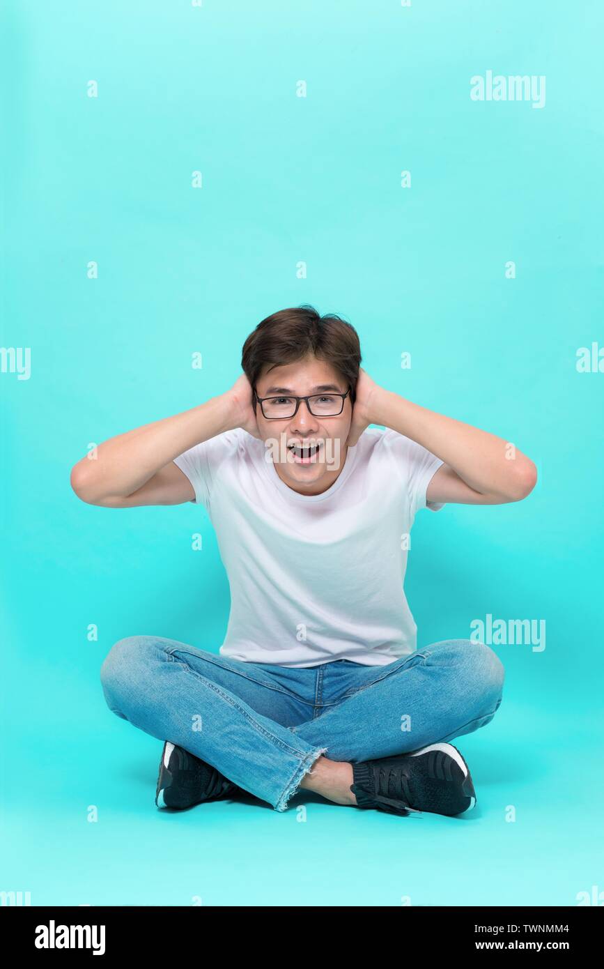 Ich bin so froh, dass Sie hier sind. Studio shot von attraktiven verspielten asiatischer Mann, Sitzen mit gekreuzten Beinen auf dem Boden, Hände auf den Kopf und Schreien von Ex Stockfoto