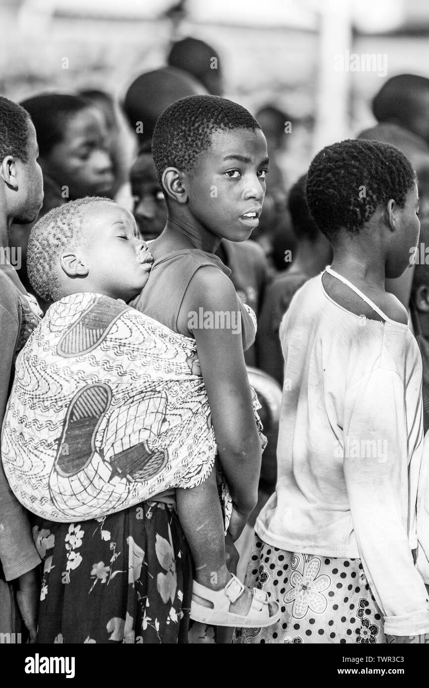 Ein Mädchen mit ihrem schlafenden Baby Schwester auf dem Rücken mit einem Stück Tuch während der Wartezeit in der Linie Essen zu bekommen Stockfoto