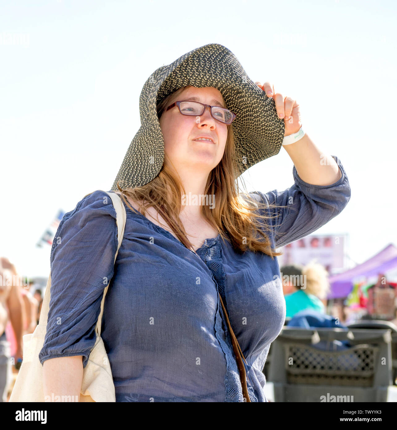 Eine junge brünette Frau in Gläser hält Ihren Sonnenhut durch die Krempe. Texas Sandfest 2019 in Port Aransas, Texas USA. Stockfoto