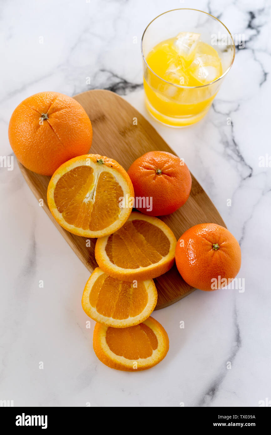 Overhead Bild des Ganzen und in Scheiben geschnittenen Orangen auf einem Holzbrett, mit einem Glas Orangensaft, alle auf einem Marmor Hintergrund Stockfoto