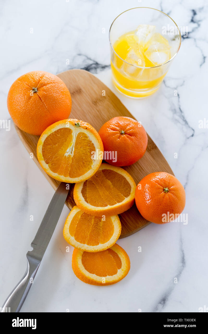 Overhead Bild des Ganzen und in Scheiben geschnittenen Orangen auf einem Holzbrett, mit einem Glas Orangensaft, alle auf einem Marmor Hintergrund Stockfoto