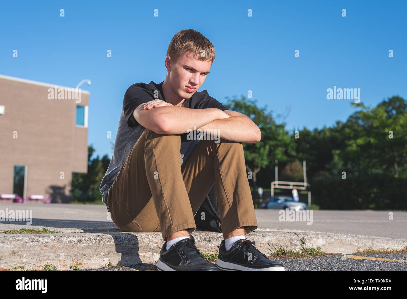 Traurige Teenager Junge sitzt auf dem Bürgersteig vor seiner Schule. Stockfoto