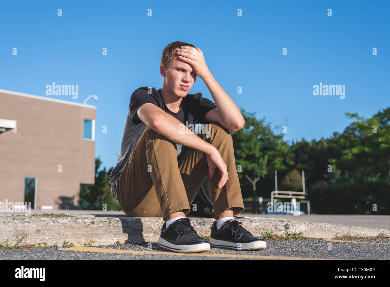 Verärgert Teenager seinen Kopf auf seine Hand, als er auf dem Bürgersteig vor seiner Schule sitzt. Stockfoto