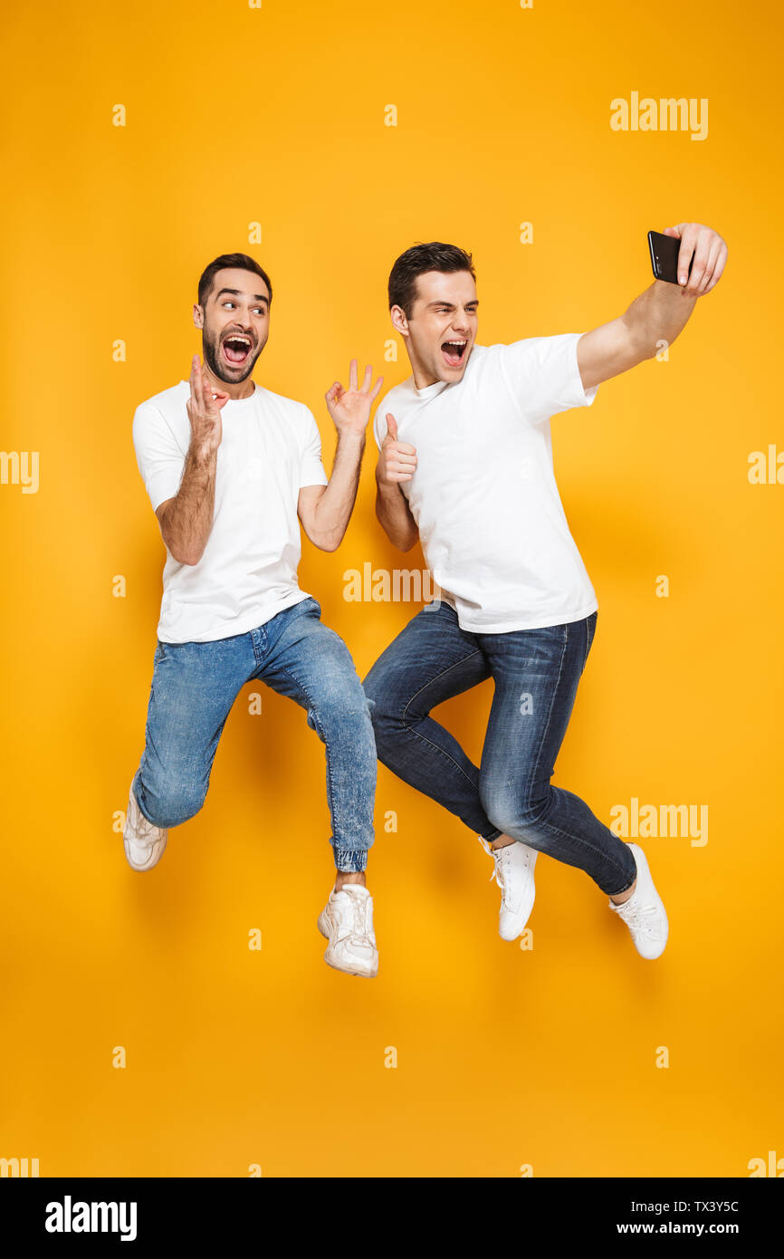 Volle Länge von zwei fröhliche aufgeregt Männer Freunde tragen Leer t-shirts über Gelb Hintergrund isoliert springen, wobei selfie Stockfoto