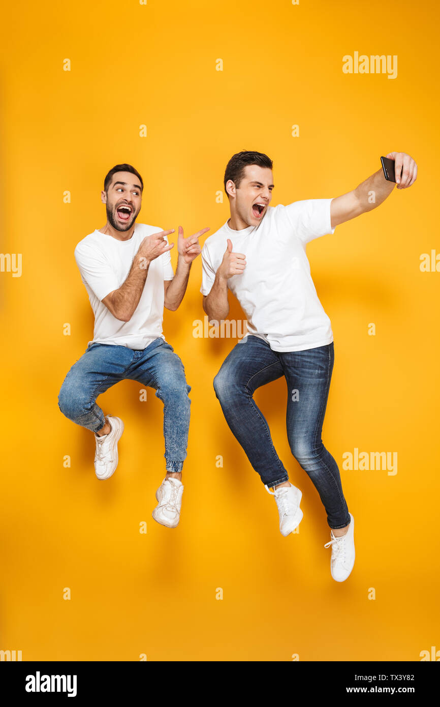 Volle Länge von zwei fröhliche aufgeregt Männer Freunde tragen Leer t-shirts über Gelb Hintergrund isoliert springen, wobei selfie Stockfoto