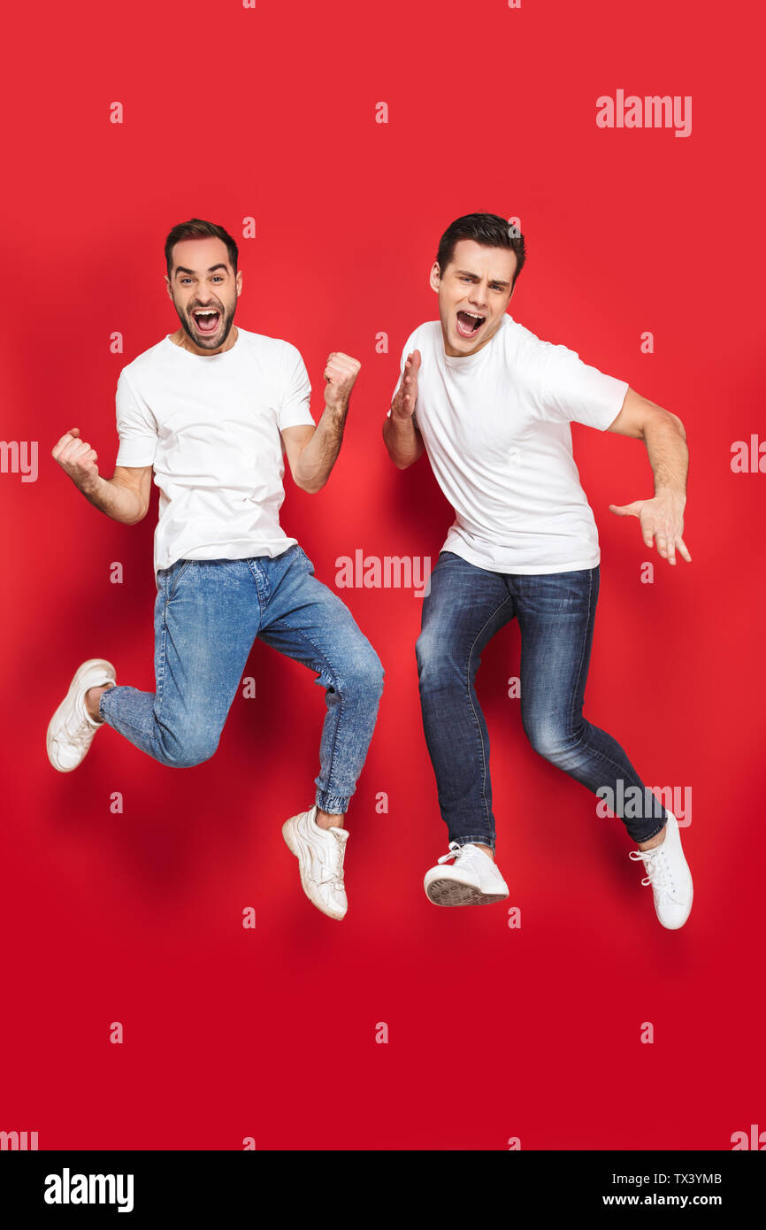 Volle Länge von zwei fröhliche aufgeregt Männer Freunde tragen Leer t-shirts über den roten Hintergrund isoliert springen, Feiern Stockfoto