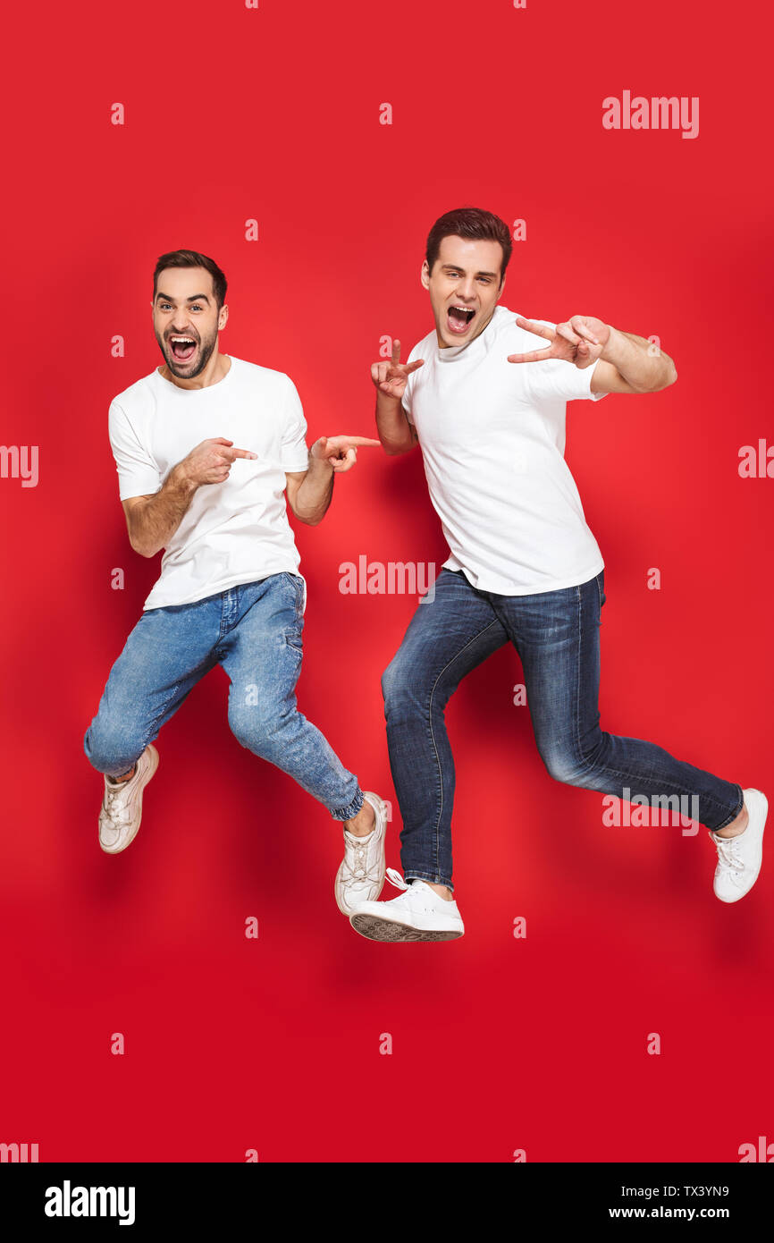 Volle Länge von zwei fröhliche aufgeregt Männer Freunde tragen Leer t-shirts über den roten Hintergrund isoliert springen, Feiern Stockfoto
