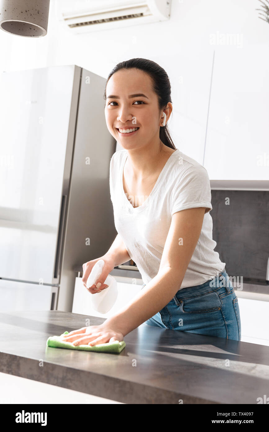 Portrait von attraktiven asiatischen Frau 20 s Tragen earpods Holding Feldspritze und Reinigung mit Reinigungsmittel und rag in der Küche zu Hause. Stockfoto