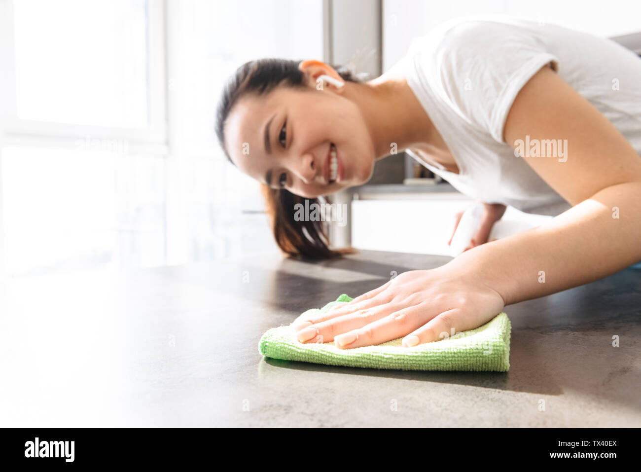 Portrait von lächelnden Asiatin 20s Tragen earpods Holding Feldspritze und Reinigung mit Reinigungsmittel und rag in der Küche zu Hause. Stockfoto