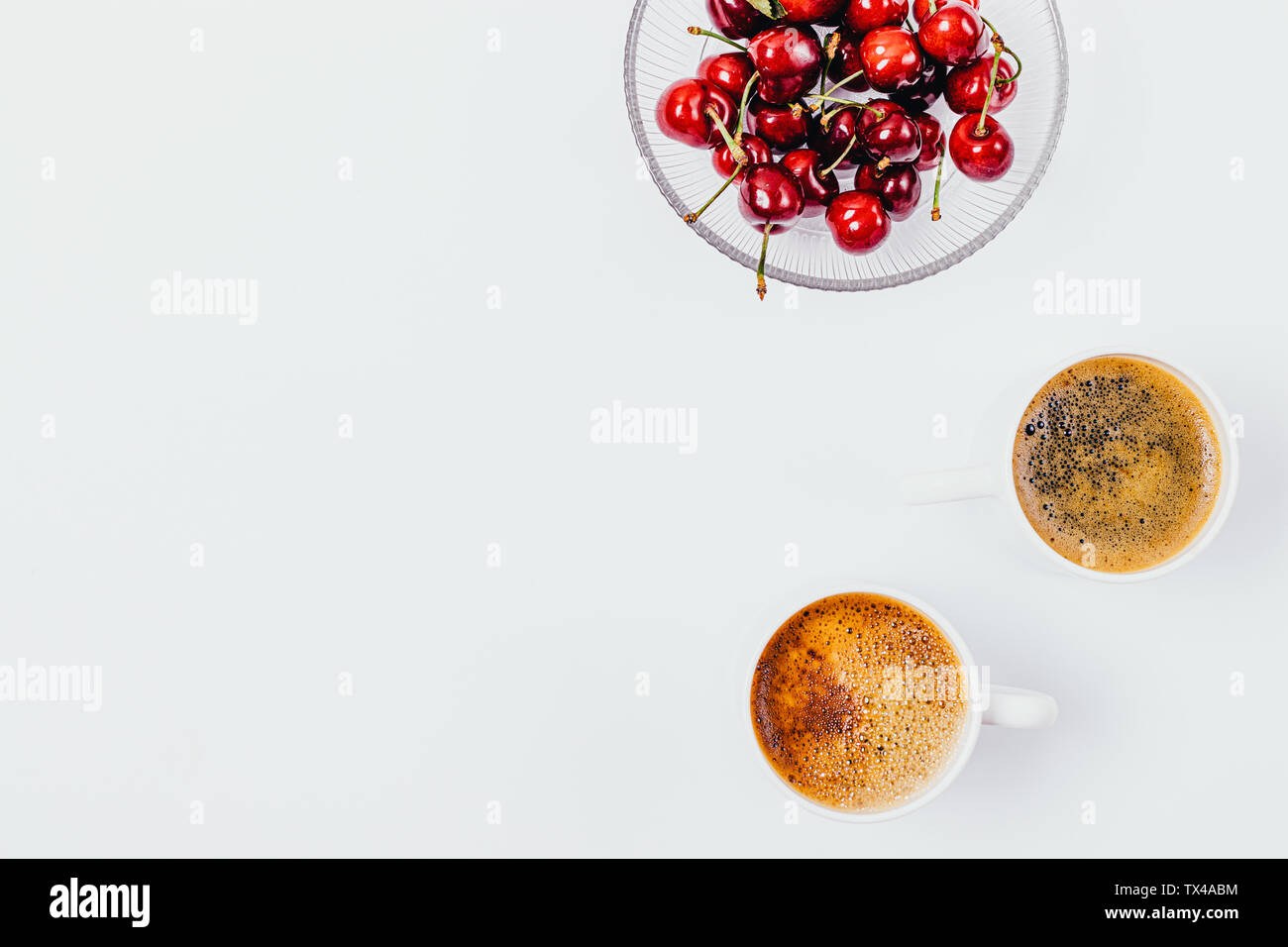 Flach Zusammensetzung der rote reife Kirschen im Glas Platte neben zwei Tassen frisch gebrühten Kaffee auf weißem Hintergrund mit kopieren. Stockfoto