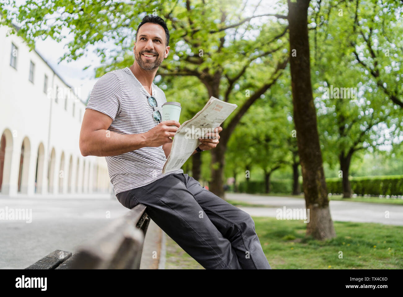 Lächelnder Mann mit Takeaway Kaffee und Zeitung auf einer Parkbank Stockfoto